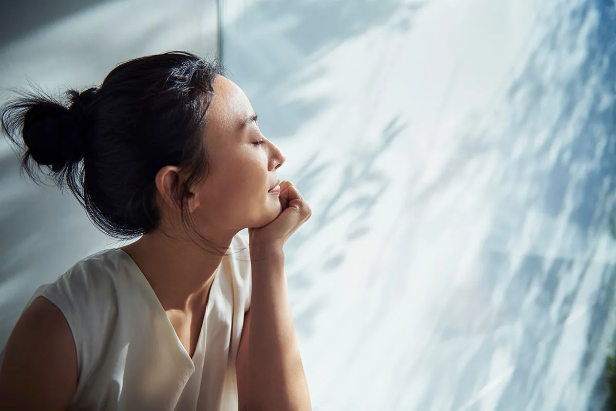 A woman sits peacefully with her eyes closed, enjoying soft natural light and a quiet moment of relaxation.