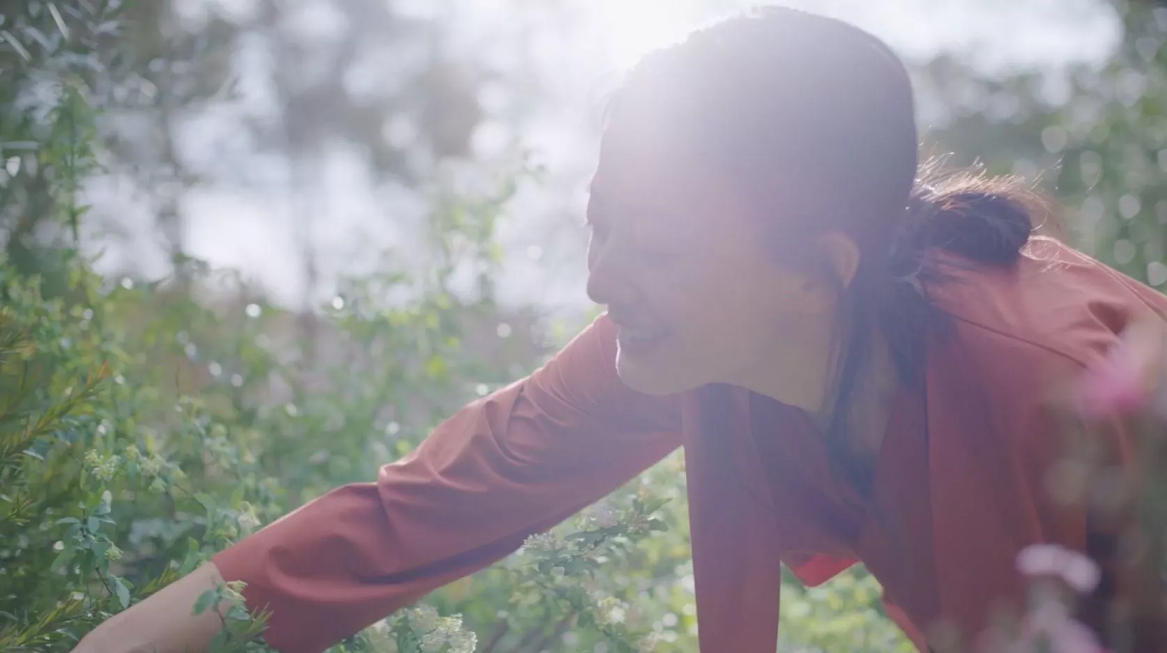 A woman smiles while tending to plants in a sunlit garden, reaching through greenery with soft light filtering from behind her.