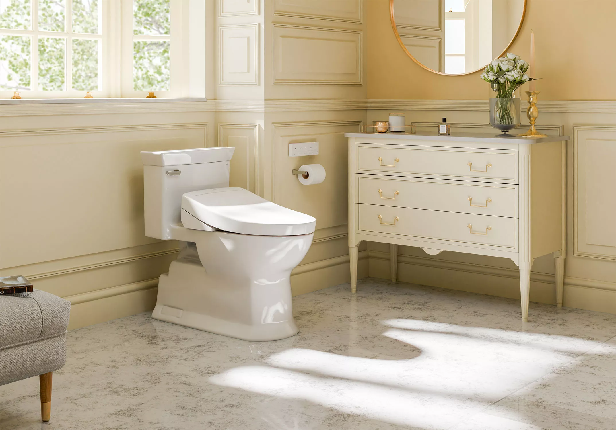 A white TOTO Aimes toilet paired with a WASHLET S5 bidet seat, installed in a bright, elegant bathroom featuring cream-colored panel walls, a marble-look floor, and a vintage-style vanity with gold hardware. Sunlight streams through a window, creating a warm, inviting atmosphere.