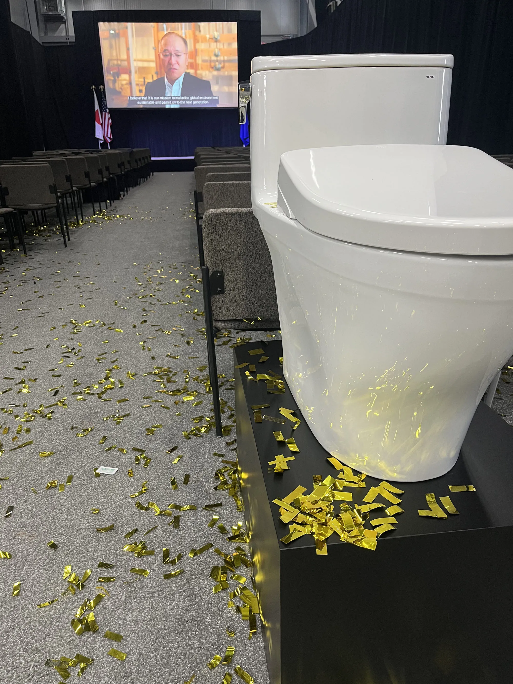 Confetti-covered aisle with a TOTO toilet display in the foreground after the Morrow ribbon-cutting, video screen in the background.
